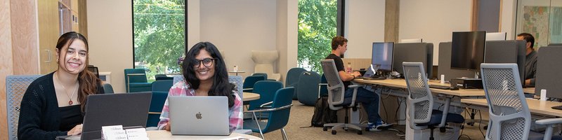 Two people smiling at the camera in a room full of comfortable seating and computers