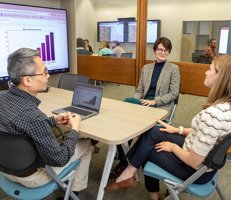 Three people meet in the foreground with a bar chart on a screen while others meet in spaces behind them