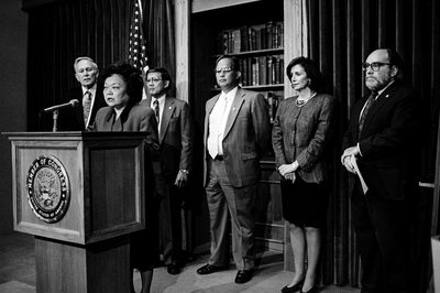 Black and white photograph of Patsy Mink speaking at a podium with 5 people behind her.