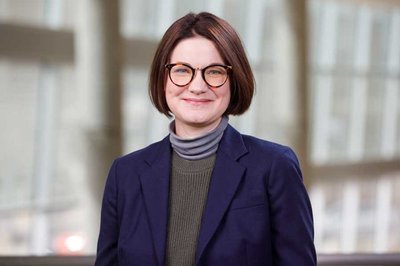 A woman with short, brown hair and glasses in a navy blue blazer and turtleneck, smiling.