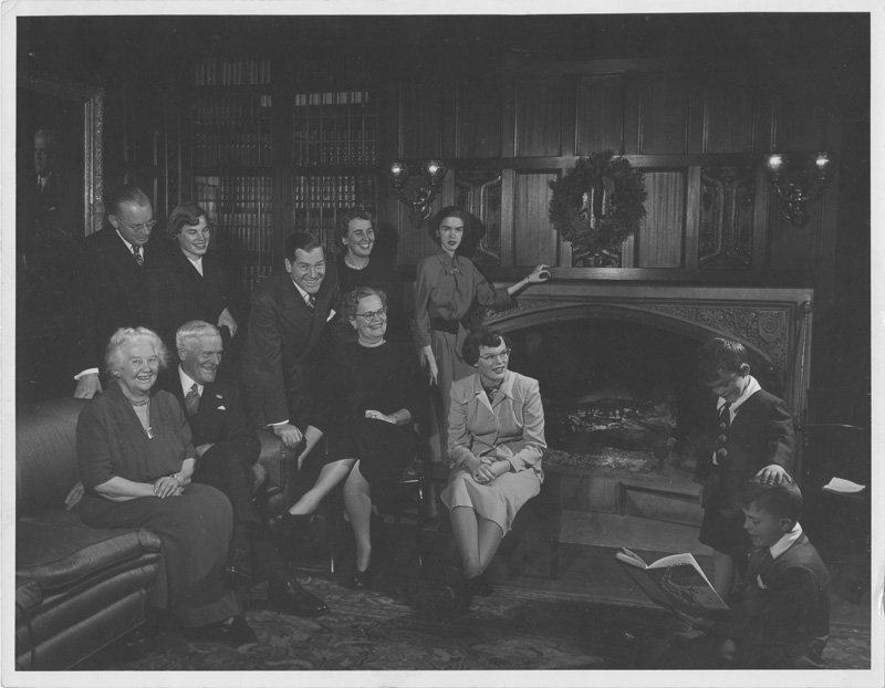 Black-and-white formal photograph of multiple generations of the Wilson family, presumably in a sitting room at Edellyn Farms. Pictured are Thomas E. Wilson, Elizabeth Foss Wilson, Helen Wilson Williams, Harry Julius Williams, Edward Foss Wilson, Pauline Wyman Wilson, three adult daughters of Helen and Harry, and two small boys of Edward Foss Wilson and Pauline Wyman Wilson. All are dressed up, and are paying attention to the two boys who are reading aloud from a book. The room is paneled in dark wood, and a Christmas wreath hangs over a large stone fireplace. There is a full bookcase to the left of the fireplace, and a painted portrait of Thomas E. Wilson hanging on the adjacent wall.